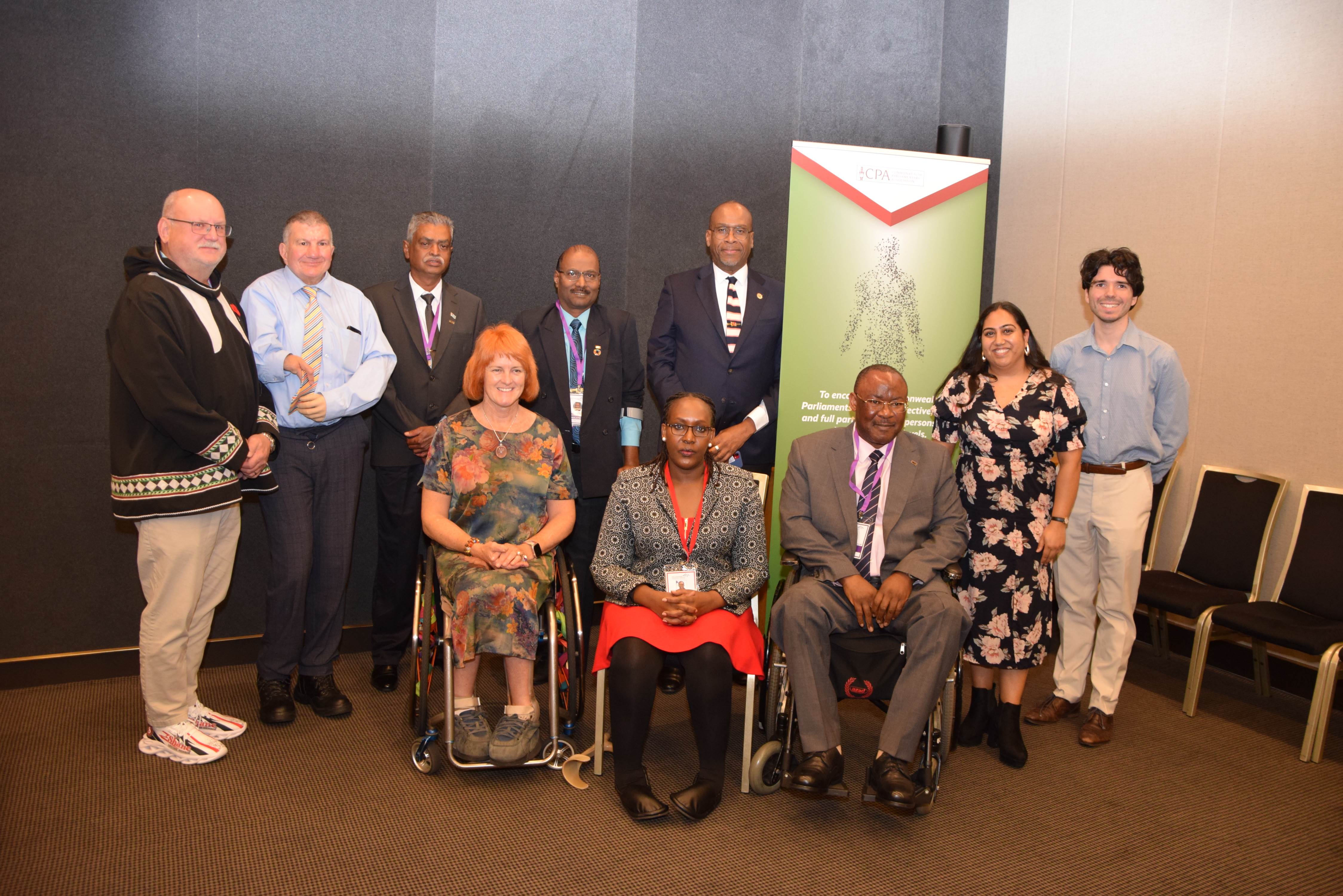 Seven people, including two wheelchair users, smile at the camera in front of banners promoting the Commonwealth Parliamentarians with Disabilities network